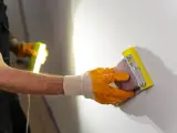 Ręka w pomarańczowej rękawicy wykonuje szlifowanie tynku gipsowego za pomocą gąbki ściernej.
