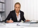 Kobieta w biurze pracuje nad dokumentami, używając kalkulatora i długopisu. Na biurku leżą stosy papierów i segregatorów, a w tle widać regał z dokumentacją. Może przygotowuje weksle.