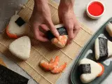 Ręce formują onigiri z krewetkami, tworząc pyszne japońskie jedzenie. Obok miska ryżu, nori i talerz gotowych przysmaków.