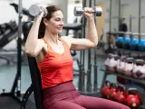 Kvinna utför axelpress med hantlar i gym. Hon sitter på en bänk och lyfter vikterna över huvudet.