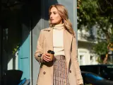 Elegancka kobieta w beżowym płaszczu, kremowym golfie i spódnicy w kratę, z kubkiem kawy w dłoni. Inspiracja na stylizacje do beżowego płaszcza.