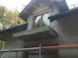 Niedokończona elewacja domu z balkonem i oknem. Na dachu siedzi ptak.