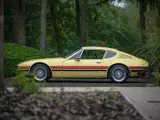 Żółty VW SP2 z czerwonymi pasami na boku, zaparkowany na tle drzew i zieleni.