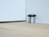 Metalowy odbojnik z czarną gumką przy ścianie, chroniący drzwi przed uderzeniem.