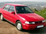Czerwony Hyundai Pony z lat 90. stoi na polnej drodze, z widokiem na dolinę i miasto w tle.