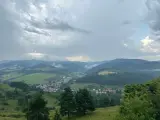 Malownicza panorama z widokiem na wieś w dolinie, otoczoną zielonymi wzgórzami i lasami. Nadciągają chmury, tworząc dramatyczne niebo nad lesnickie sedlo.