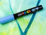 En ljusblå Posca-tuschpenna ligger på en abstrakt målning i gröna och blå nyanser. Perfekt för att måla med tuschpennor.