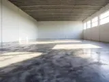 Pusta hala z betonową podłogą, która lśni jak polerowana żywica. Światło wpada przez wysokie okna, tworząc na niej refleksy.