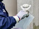 Pracownik w niebieskiej kurtce wykonuje tynkowanie elewacji, nakładając zaprawę kielnią na ścianę budynku.