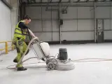Pracownik w kamizelce odblaskowej wykonuje szlifowanie betonu za pomocą maszyny.