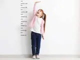 Dziewczynka z długimi włosami w kucykach, ubrana w różowy sweterek i jeansy, z radością wyciąga rękę do góry przy miarce wzrostu. To wizualizacja tego, jak pobudzić wzrost u dziecka.