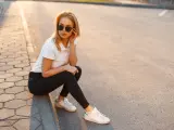 Blondynka w czarnych jeansach i białej koszulce polo, siedząca na chodniku. Stylowa inspiracja, jak nosić koszulka polo damska na co dzień.