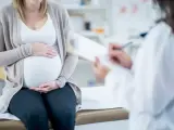Kobieta w ciąży siedzi na kozetce, trzymając dłonie na brzuchu. Lekarz spisuje dokumenty, prawdopodobnie wystawiając zaświadczenie o ciąży.