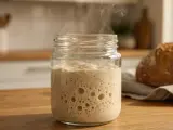 Blubbernder Sauerteig im Glas, der nach Essig riecht, dampft leicht. Daneben liegt ein frisch gebackenes Brot.
