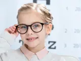 Uśmiechnięta dziewczynka w okularach, z tablicą optometryczną w tle. Okulary nosi się od jakiej wady, by widzieć wyraźnie.
