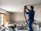Hombre lijando techo con gotelé para tapar con pladur. El salón está cubierto con plásticos protectores.
