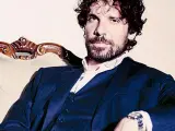 L'attore Montanari, con barba e capelli ricci, indossa un completo blu e una camicia bianca, seduto su una poltrona d'epoca.