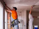 Deux ouvriers rénovent un bâtiment, l'un ajuste des câbles près d'une fenêtre, l'autre enduit le plafond. Ces travaux sont essentiels pour les aides aux collectivités locales.