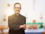 Ein lächelnder evangelischer Pfarrer mit Brille und überkreuzten Armen steht vor einem unscharfen Kircheninterieur.