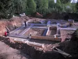 Budowa fundamentów: zbrojenie i szalunki czekają na chudy beton. W tle las i plac budowy.