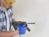 Man in safety glasses and blue gloves holding a drill to a white surface.