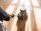 En person slipar ett trägolv med en maskin. Dammet virvlar upp och skapar ett mönster på det ljusa träet.