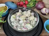 Pyszna sałatka z młodych ziemniaków z czerwoną cebulą i koperkiem, podana w szklanej misie. Obok rzodkiewki i ogórki kiszone.