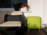 Zielony kompostownik na balkonie, obok wygodnej kanapy z poduszką w grochy.