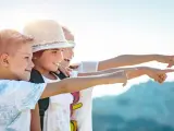Trois enfants souriants, portant des sacs à dos, pointent du doigt vers la droite, sous un ciel ensoleillé.