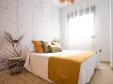 Habitación con papel tapiz de ramas y gotele moderno. Cama con cojines verdes y naranjas, y manta mostaza.