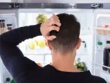Un homme se gratte la tête devant un réfrigérateur ouvert, plein de nourriture. Il semble réfléchir à un réglage frigo.