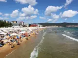 Tłum ludzi wypoczywa na piaszczystej plaży Słoneczny Brzeg, opalając się na leżakach pod białymi parasolami. W tle widać morze i zabudowania kurortu.