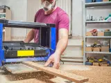 Stolarz w masce ochronnej pracuje przy strugarce, wygładzając drewno. Wióry sypią się wokół.