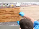 Mani guantate applicano un trattamento per come indurire il legno. Tavolo in legno con diverse lattine di finitura.