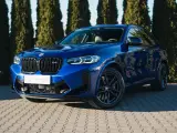 Błyszczący, niebieski suv BMW X4 M Competition zaparkowany przed żywopłotem.