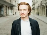 Bartosz Bednarski na tle miejskiej ulicy, z rudymi włosami i zielonymi oczami, w białej koszuli i ciemnej kurtce.