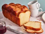 Ein goldbraunes Brioche Brot Rezept, aufgeschnitten auf einem Holzbrett mit Marmelade und Tassen im Hintergrund.