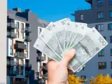 Dłoń trzyma banknoty 100 zł na tle nowoczesnych budynków mieszkalnych. Symbolizuje to podatek od sprzedaży nieruchomości.
