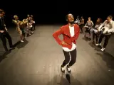 Un danseur afro 93, en veste rouge, mène une troupe sur scène. Les danseurs, aux tenues variées, exécutent des mouvements synchronisés.
