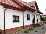 Biały dom z brązowymi cokołami, czerwonym dachem i ozdobnymi schodami.
