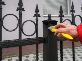 Ręka w czerwonej kurtce maluje pędzlem czarną, kutą balustradę balkonu w bloku.