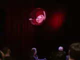 Na scenie, wśród czerwonych zasłon, unosi się kula z projekcją twarzy, przypominająca kurkę wodną. Widownia wpatruje się w to widowisko.