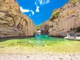 Spiaggia di ciottoli e acque cristalline tra le rocce dell'isola di Lissa, con persone che nuotano e barche.