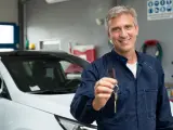 Uśmiechnięty mechanik w granatowym kombinezonie podaje kluczyki do białego samochodu. W tle warsztat samochodowy, gdzie wszystko jest gotowe, by sprawdzić, co to homologacja.