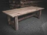 Ett rustikt bord av oljad ek med grova ben och en bred bordsskiva. Bakom syns staplade trästockar.