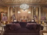 Salón lujoso con sofás de terciopelo morado, lámparas doradas y un candelabro de cristal. El techo y las paredes doradas evocan el estilo barroco español.