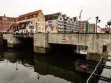 Kamienny most nad ciemną wodą, z rzędem zabytkowych kamienic w tle. W oddali widać dźwigi budowlane.