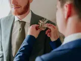 Un homme ajuste une boutonnière sur le costume d'un autre. Divers types de boutonnières existent, celle-ci est composée de fleurs séchées et de feuillages dorés.