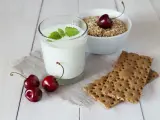 Un verre de yaourt lait végétal, garni de menthe, avec des cerises et des crackers sur une table en bois.