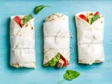 Trzy keto tortille z siemienia lnianego, zawinięte w papier i przewiązane sznurkiem, z widocznym nadzieniem z kurczaka, pomidorów i szpinaku.